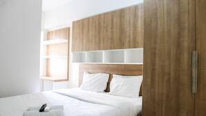 A modern hotel room with a wooden headboard, white bedding, and a remote control on the bed. - 2BR with Sofa Bed at Springlake Summarecon Apartment (Bekasi)