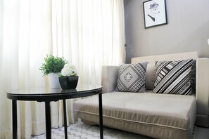 A living room with a beige sofa, patterned pillows, a small round table, a potted plant, and a framed picture on the wall.