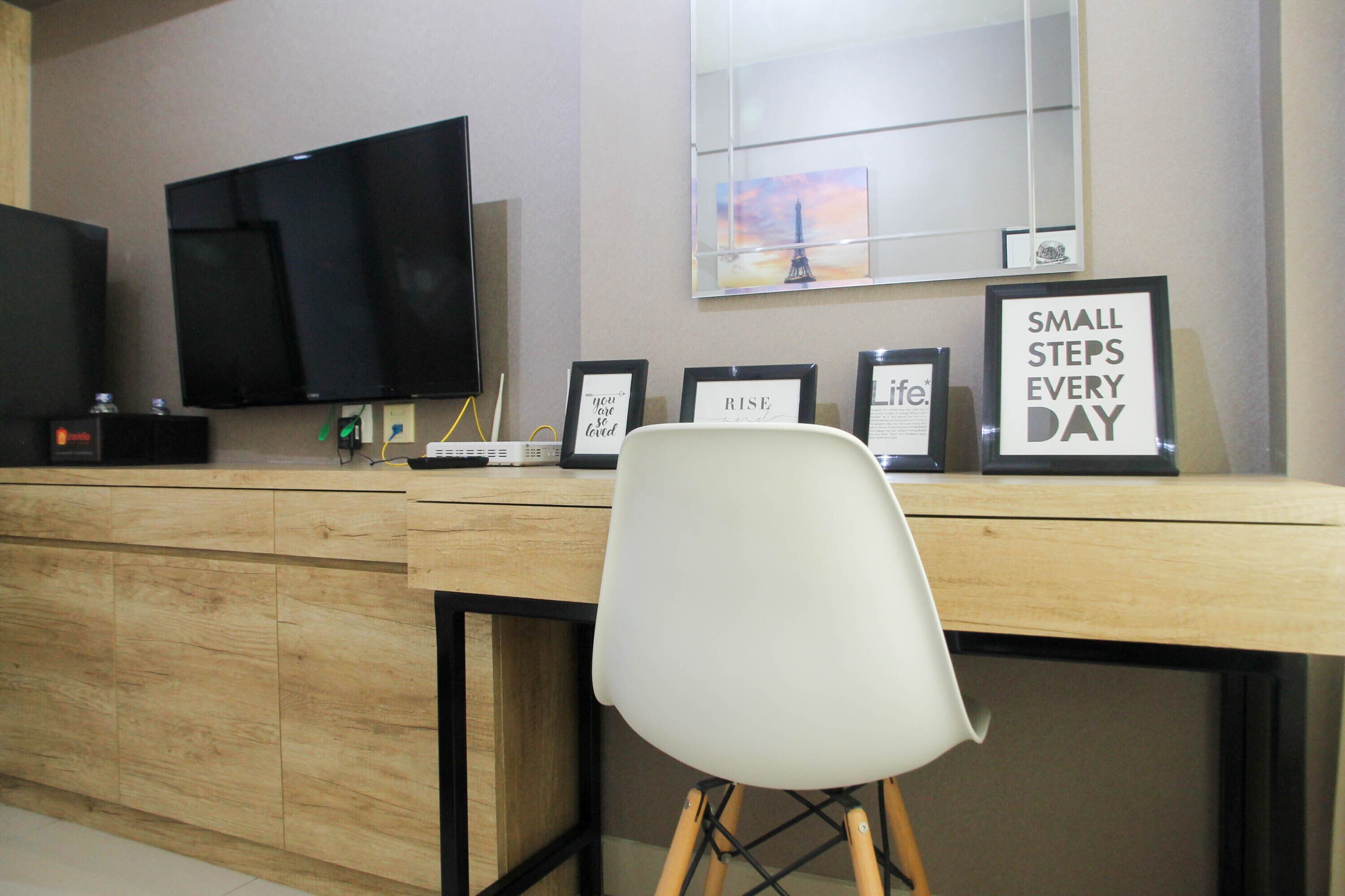 Een kamer met een houten bureau, een witte stoel, een flatscreen televisie en gemotiveerde citaten in lijsten aan de muur.