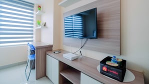 A modern hotel room with a flat-screen TV mounted on the wall, a desk with a computer and a remote control, a blue chair, and a minibar with travelio branding. - Wonderful Studio The Springlake Summarecon Apartment (Bekasi)