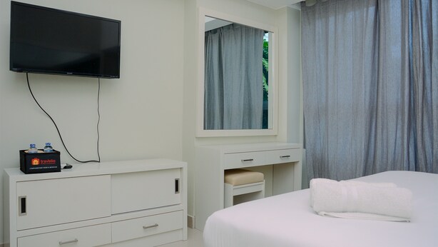 A hotel room with a flat-screen TV mounted on the wall, a white dresser with drawers, a bedside table with a towel, and a window with curtains. - Studio Apartment with Garden View at The Springlake View Summarecon (Bekasi)