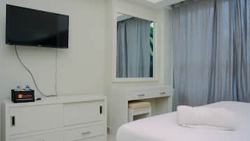 A hotel room with a flat-screen TV mounted on the wall, a white dresser with drawers, a bedside table with a towel, and a window with curtains.