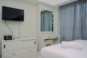 A hotel room with a flat-screen TV mounted on the wall, a white dresser with drawers, a bedside table with a towel, and a window with curtains.
