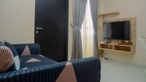 Un salon avec un canapé de couleur sombre, un téléviseur à écran plat fixé sur une étagère en bois et une porte dotée d’une poignée.