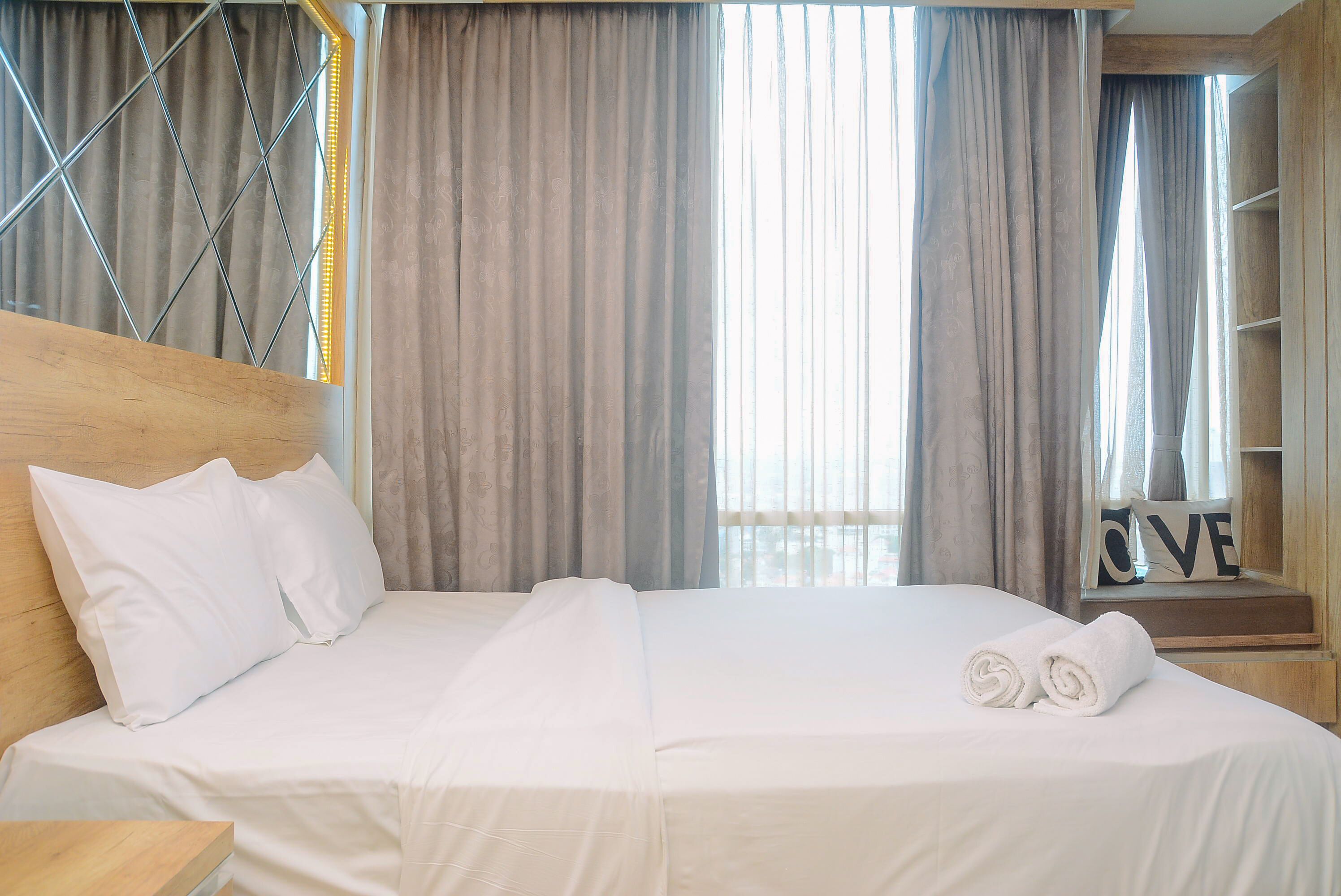 Una cama bien tendida con sábanas blancas y dos toallas enrolladas, un cabecero de madera y un ventanal con cortinas.