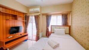A hotel room with a bed, a TV on a wooden stand, a window with curtains, and an air conditioning unit.