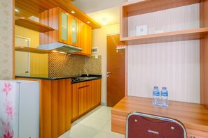 A compact kitchen with wooden cabinets, a tiled backsplash, and a built-in oven. There is a refrigerator with a floral design, a water dispenser, and a wooden shelf with two water bottles.