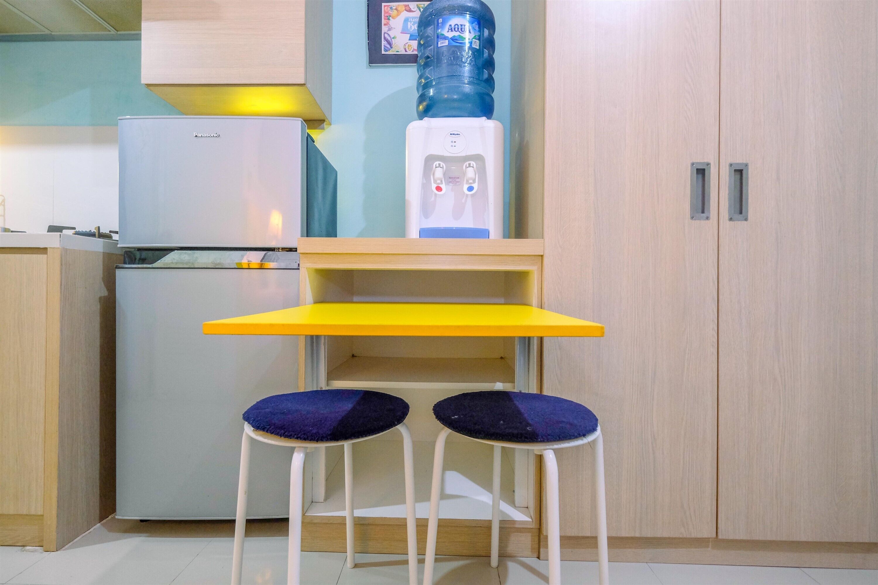 Una cocina compacta con refrigerador, dispensador de agua y dos taburetes con cojines azules.