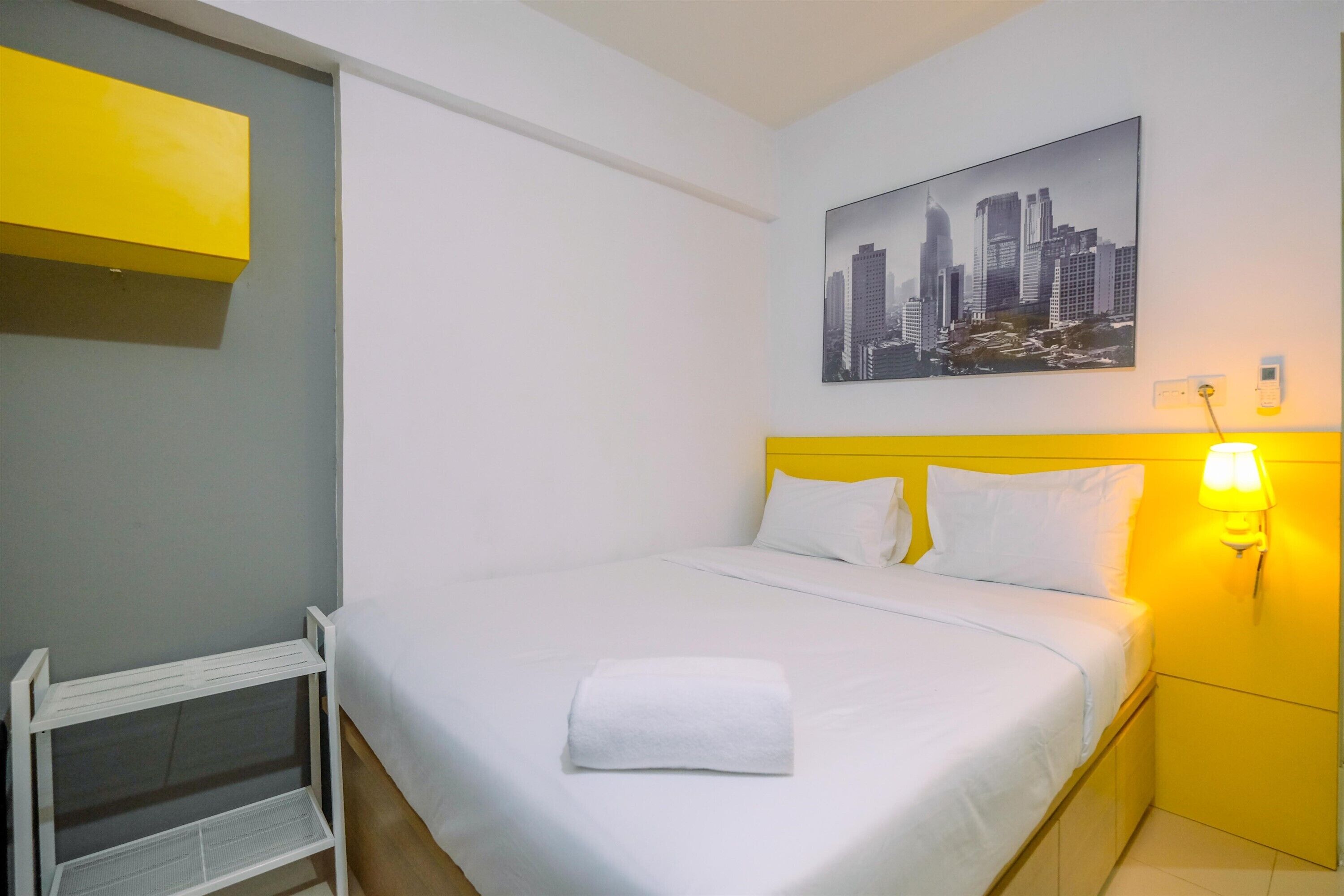 Une chambre d’hôtel avec un lit, un tableau représentant un paysage urbain, une table de chevet avec une lampe et une petite étagère.