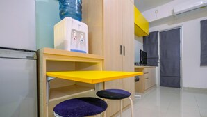 A compact room with a kitchenette, a water dispenser, a yellow table, two purple cushioned stools, and a closed door. - Warm and Cozy Studio Park View Condominium Apartment (Depok)