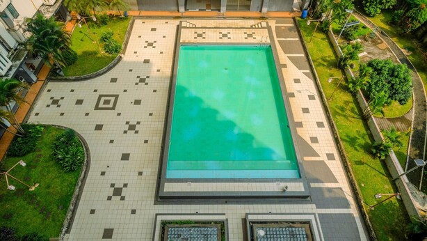 An aerial view of a swimming pool surrounded by a tiled deck with a geometric pattern, bordered by greenery and a low wall.