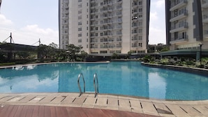 A swimming pool with a wooden deck and metal ladders, surrounded by greenery and modern buildings. - Compact and Modern Studio Room at Casa De Parco Apartment (Pagedangan)