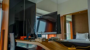 A modern hotel room with a large flat-screen TV mounted on a wooden wall, a desk with a chair, and a bed with a white pillow. - Compact and Modern Studio Room at Casa De Parco Apartment (Pagedangan)