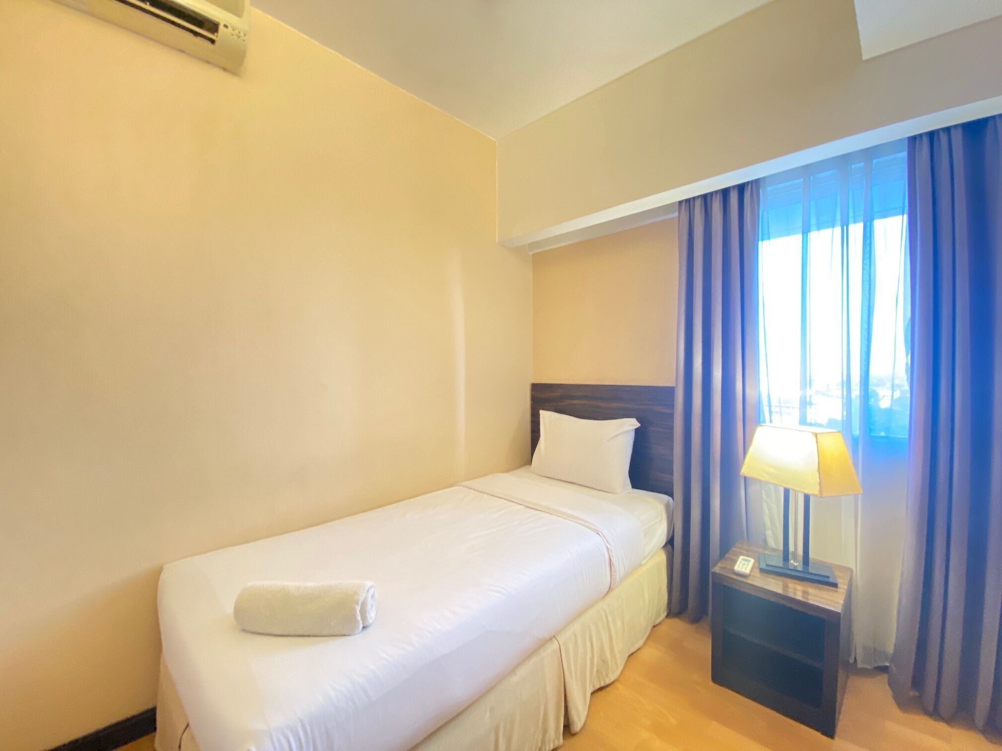 A single bed with a white mattress and pillow, a bedside table with a lamp, and a window with blue curtains.
