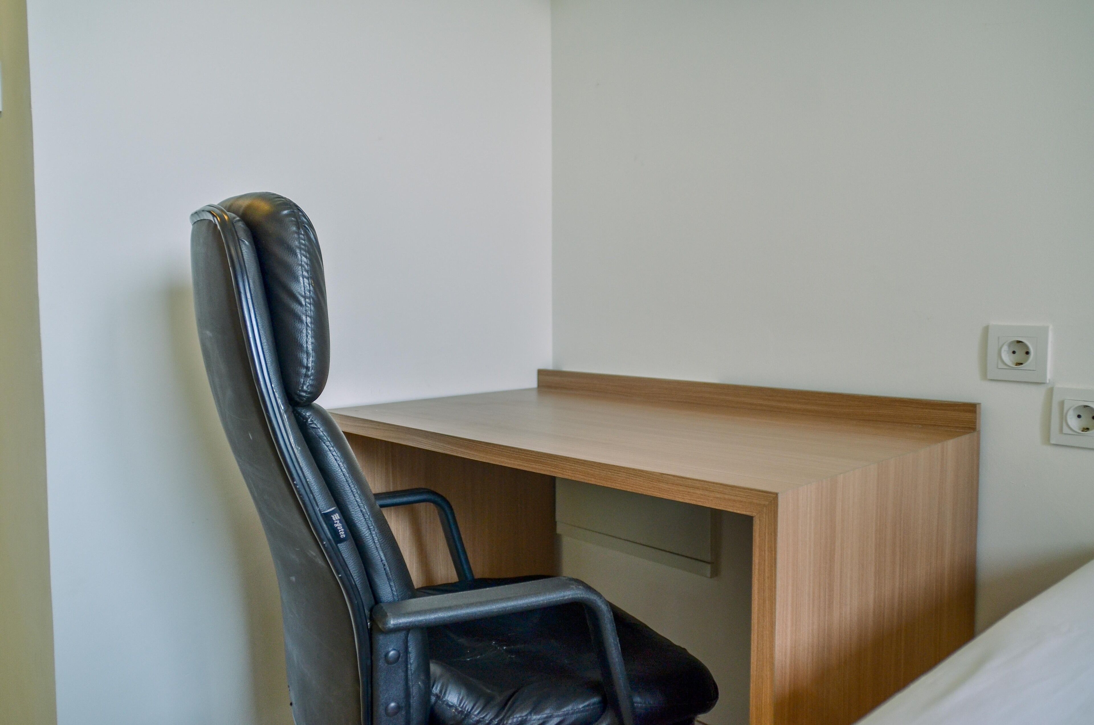 Ein Büro im Eck mit einem Holztisch, einem schwarzen Ledersessel und einer weißen Wand mit Steckdose.