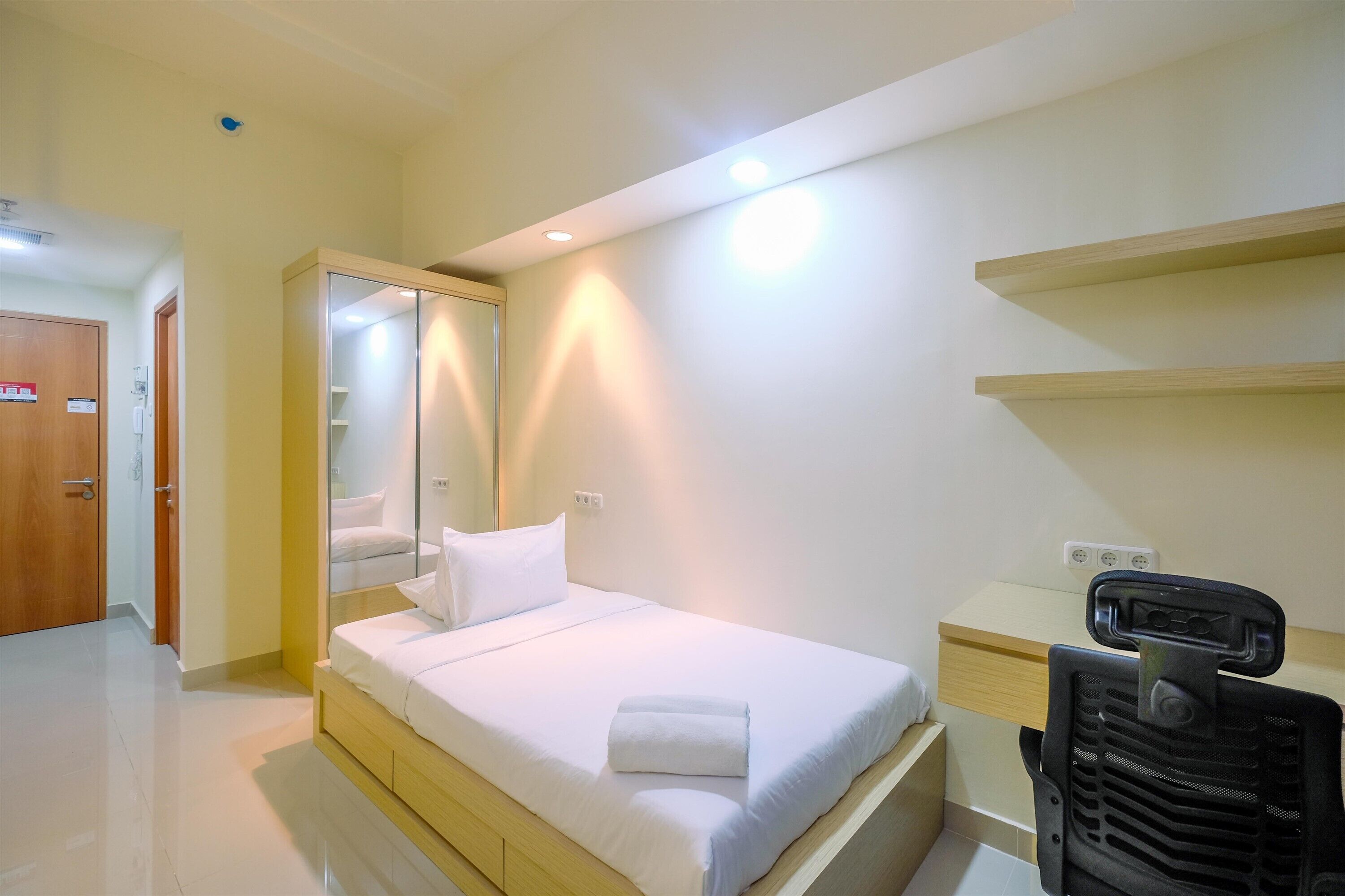 A hotel room with a bed, a desk, a chair, a mirror, and a shelf.