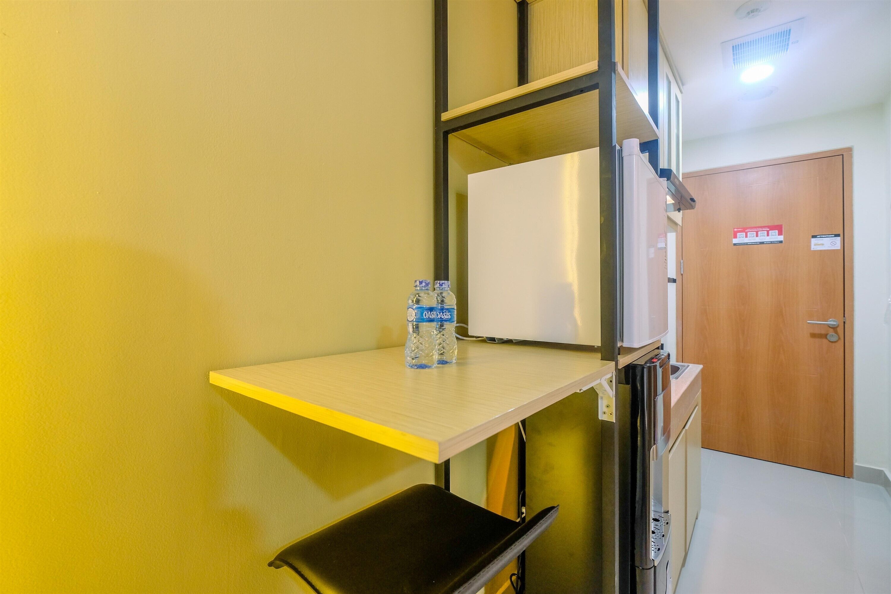 Une chambre d’hôtel équipée d’une kitchenette, d’une petite table avec une bouteille d’eau et d’une porte sur laquelle est apposée une pancarte.