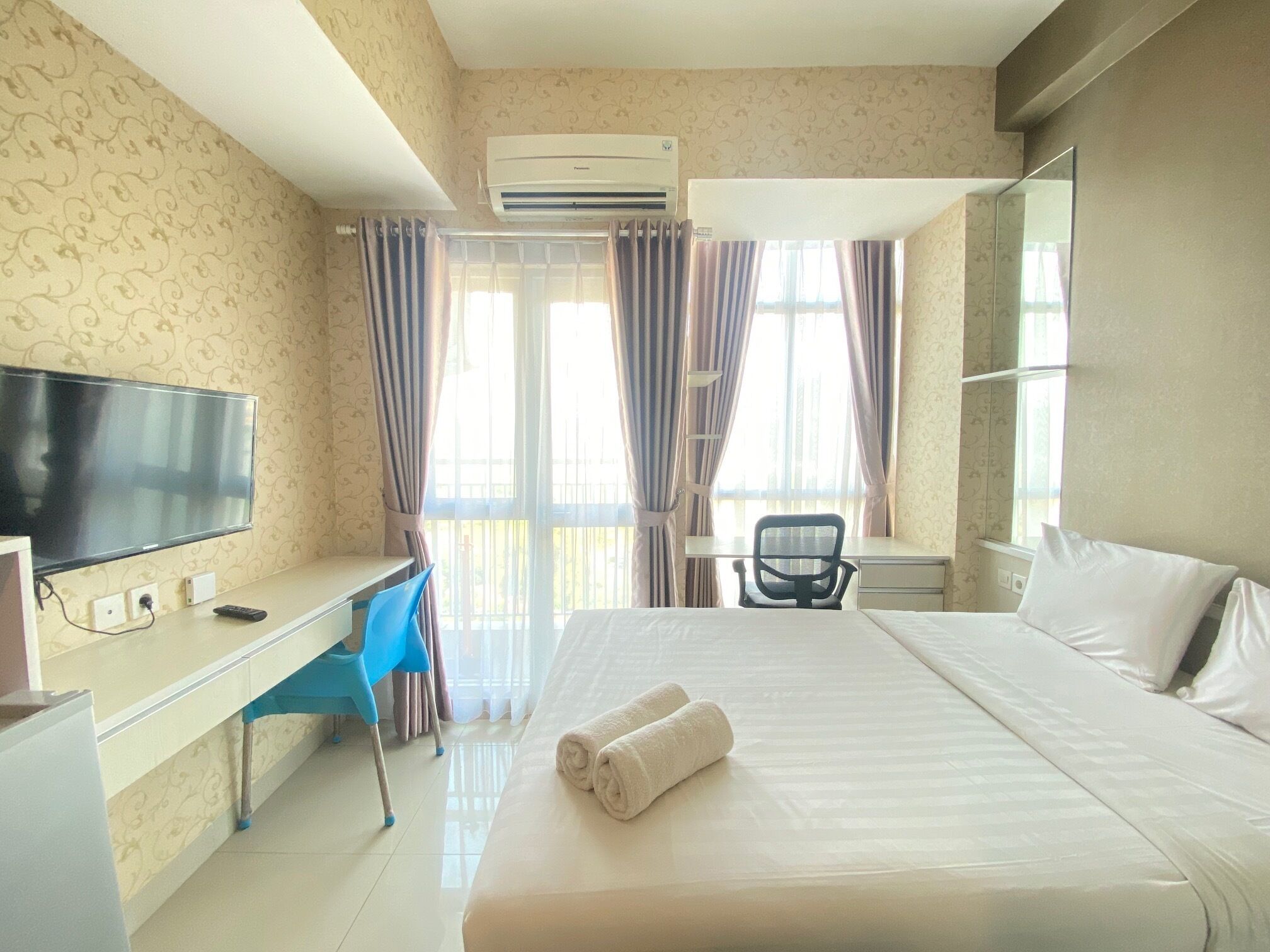 A hotel room with a bed, a desk, a chair, a TV, and an air conditioning unit.