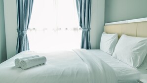 A neatly made bed with white linens and two rolled towels, a beige headboard, and light blue curtains. - Homey and Comfy 1BR at Casa de Parco Apartment (Pagedangan)