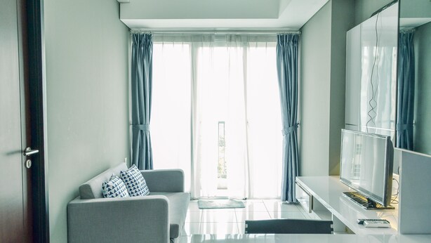 A modern living room with a grey sofa, a flat-screen TV, and a dining area with a white table. - Homey and Comfy 1BR at Casa de Parco Apartment (Pagedangan)