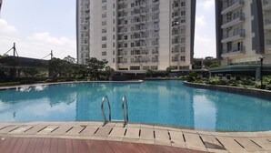A swimming pool with a wooden deck and metal ladders, surrounded by greenery and modern buildings. - Homey and Comfy 1BR at Casa de Parco Apartment (Pagedangan)
