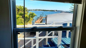 Une vue à travers une fenêtre sur une terrasse avec une rambarde en bois, un plancher bleu et une jetée qui s’avance dans une étendue d’eau.