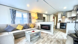 A modern living room with a sofa, coffee table, and fireplace. The kitchen area includes a microwave, oven, and cabinets. - 63 Bude Perran Paradise (Newquay)