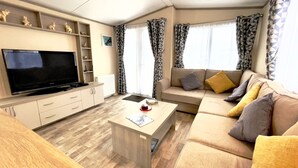 A modern living room with a flat-screen TV, a sofa with cushions, a coffee table, and a bookshelf. - 63 Bude Perran Paradise (Newquay)