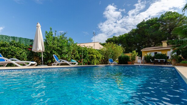 A clear blue swimming pool with lounge chairs and umbrellas, surrounded by lush greenery and a building in the background.