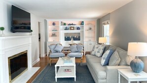 Ein Wohnzimmer mit Kamin, zwei Sesseln, einer Couch und einem Couchtisch, sowie einem an der Wand befestigten Fernseher.