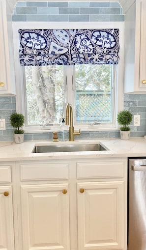 Kitchen features a central sink, cabinetry with gold handles, and a window above with a valance.