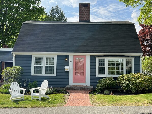 A two-story house with a dark blue exterior, a red door, and a chimney. There are two white windows and a small porch with a brick pathway leading to the door.