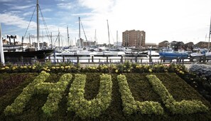 Un port de plaisance avec des bateaux amarrés, un bâtiment en arrière-plan et un jardin où une haie forme les lettres « HULU ».