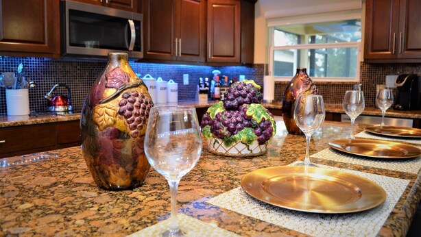 A kitchen with granite countertops, wooden cabinets, and a backsplash. There are wine glasses, a bowl of grapes, and a decorative vase on the counter. - Exquisite Destination Villas... Crystal location (Kissimmee)