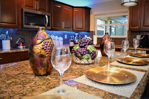 A kitchen with granite countertops, wooden cabinets, and a backsplash. There are wine glasses, a bowl of grapes, and a decorative vase on the counter.