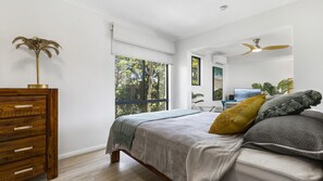 Relax in a serene space with a view of lush greenery, featuring a comfortable bed, dresser, and flat-screen TV.