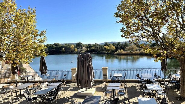 An outdoor seating area with tables and chairs facing a lake, surrounded by trees and a railing. - Apartment 4/5 persons Domaine et Golf international de Pont Royal (Mallemort)