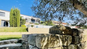 A stone wall with a small water feature, a tall green tree, and a building with a white facade in the background. - Villa Loralil by Costabravaway (calonge)