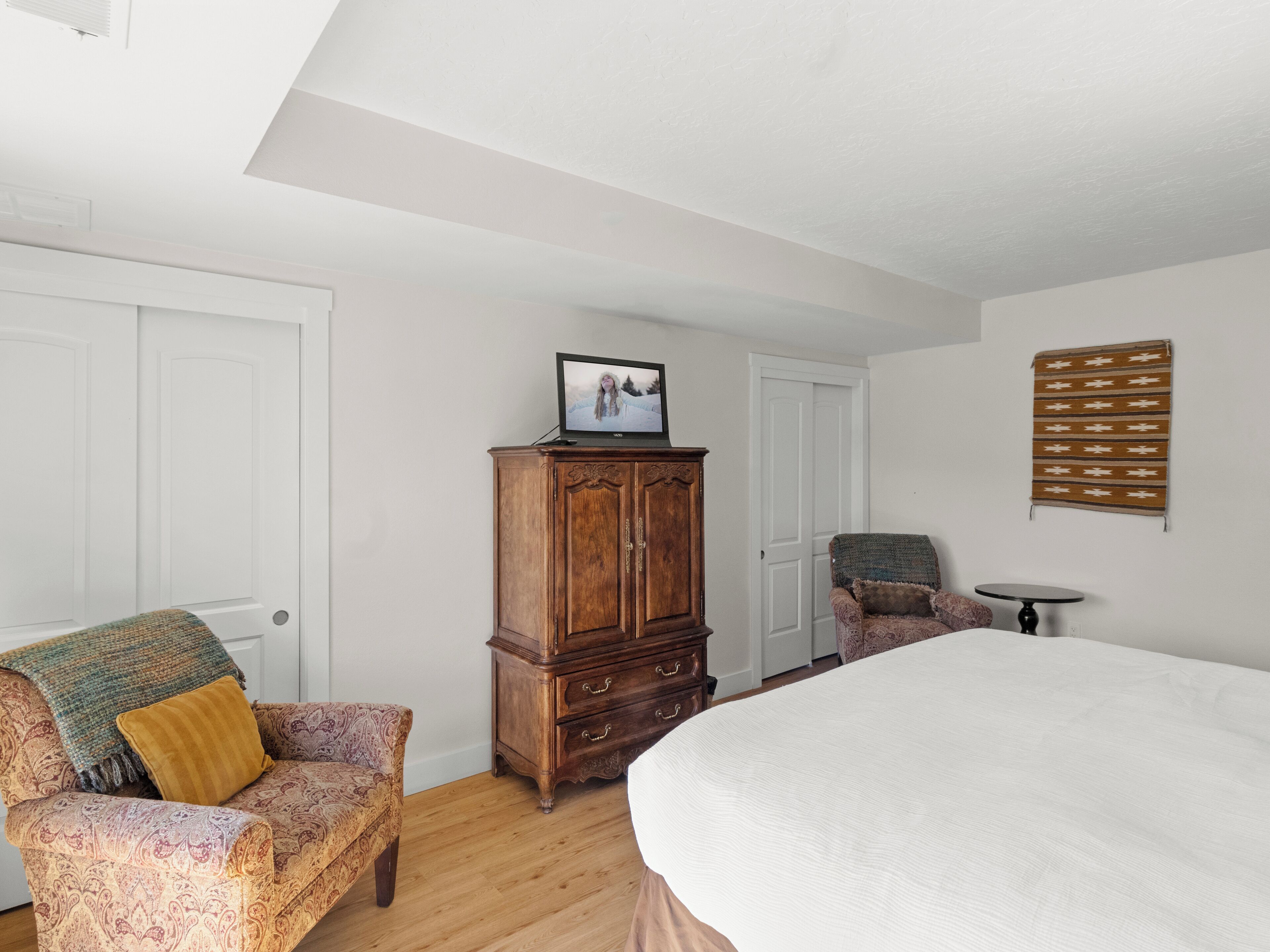 Une chambre à coucher avec un grand lit, une armoire en bois, un fauteuil à motifs, une petite table et une tapisserie murale.