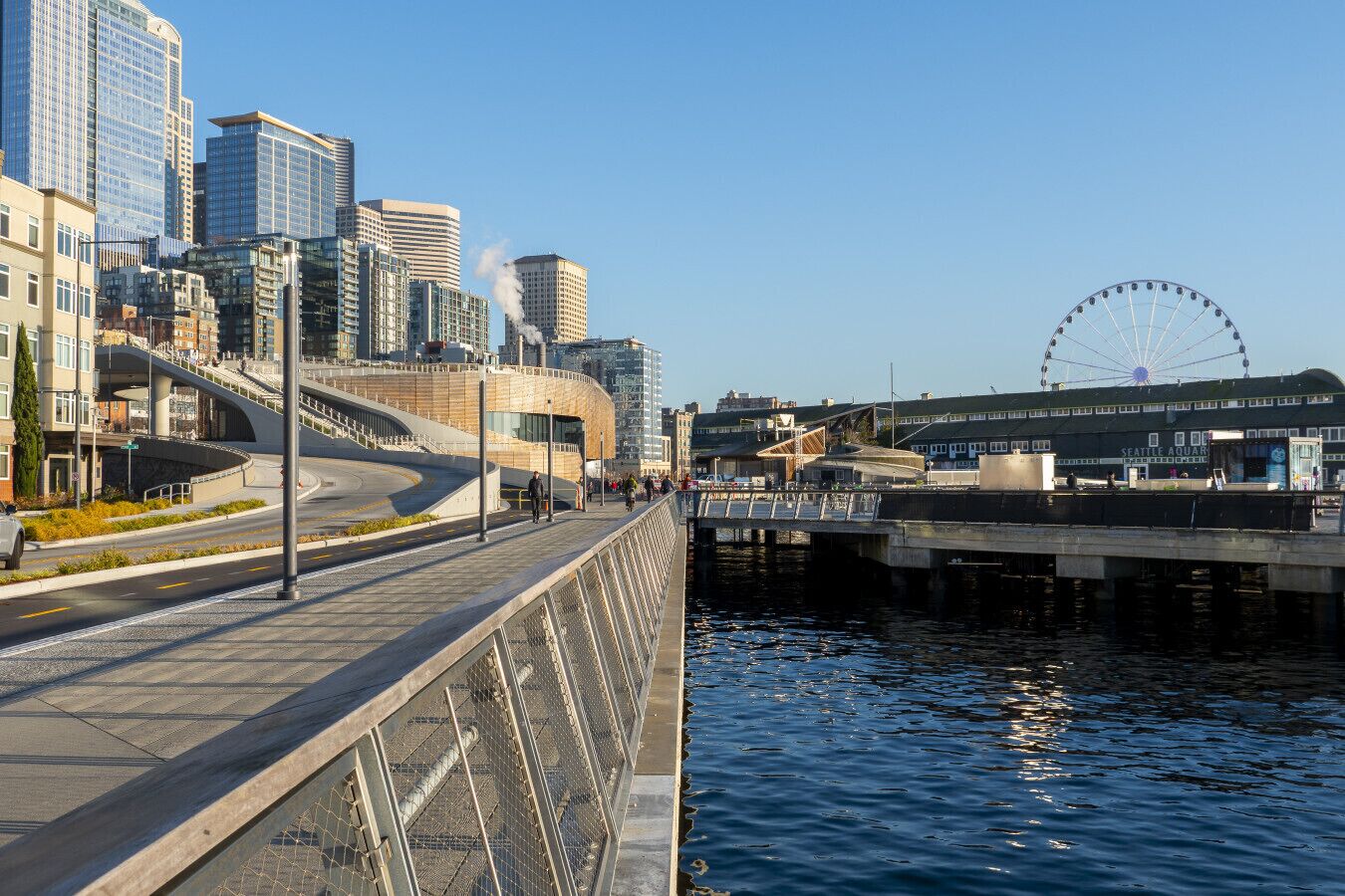 A waterfront promenade with modern buildings, a Ferris wheel, and a bridge.