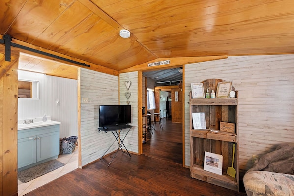 Interior acogedor de una cabaña con techo de madera, un televisor, un estante de madera con varios objetos y un letrero que dice 'Welcome'.