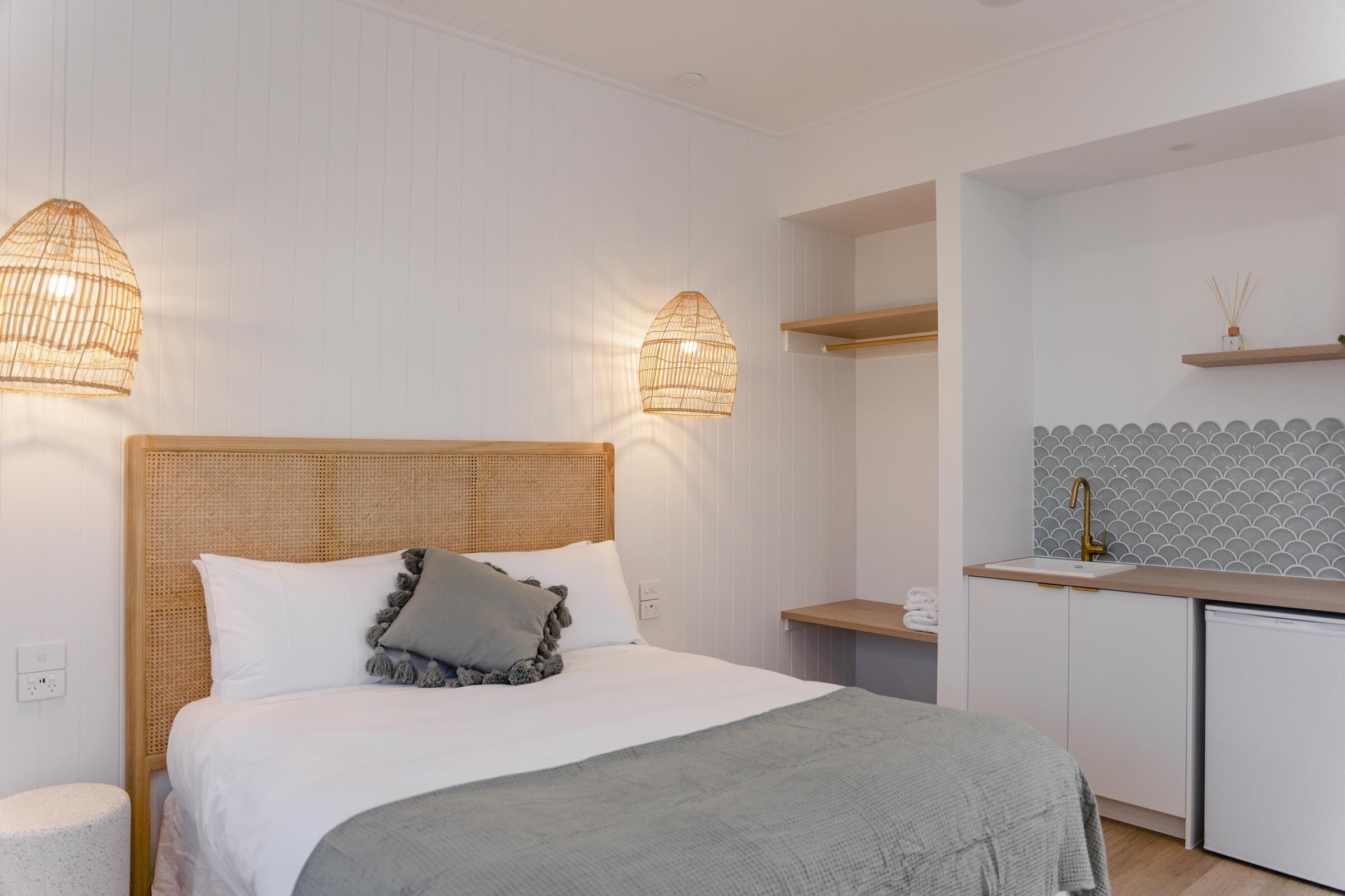 Une chambre à coucher comprenant un lit, des lampes de chevet, une table de chevet, une petite étagère et une kitchenette équipée d’un évier et d’un plan de travail.