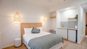 Una camera da letto con un letto, lampade da comodino, uno sgabello rotondo e un angolo cottura con lavello e una pianta.