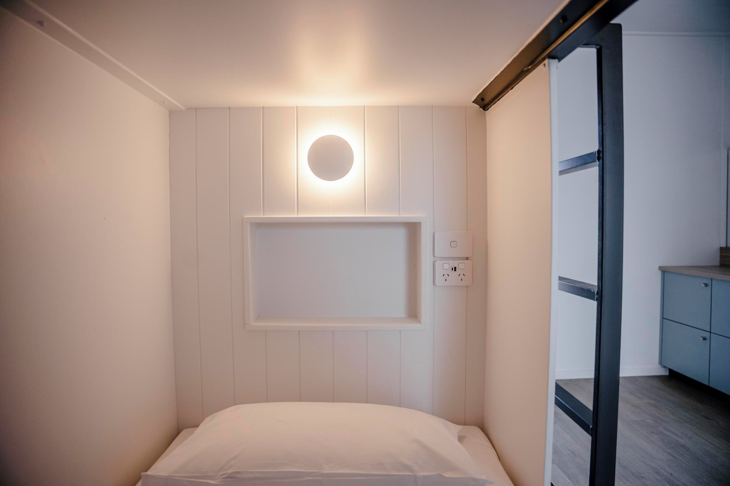 Una habitación pequeña y privada con cama, luz empotrada en la pared y toma de corriente.