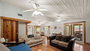 Uma sala de estar espaçosa com piso de madeira, dois sofás, ventiladores de teto e uma área de refeições visível através de portas de vidro.