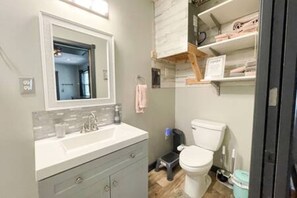A bathroom with a white sink, mirror, and a toilet. There is a shelf with folded towels and a trash bin. - 5 Tiny Town + Monthly + Utilities + Comfy Stay (Hico)