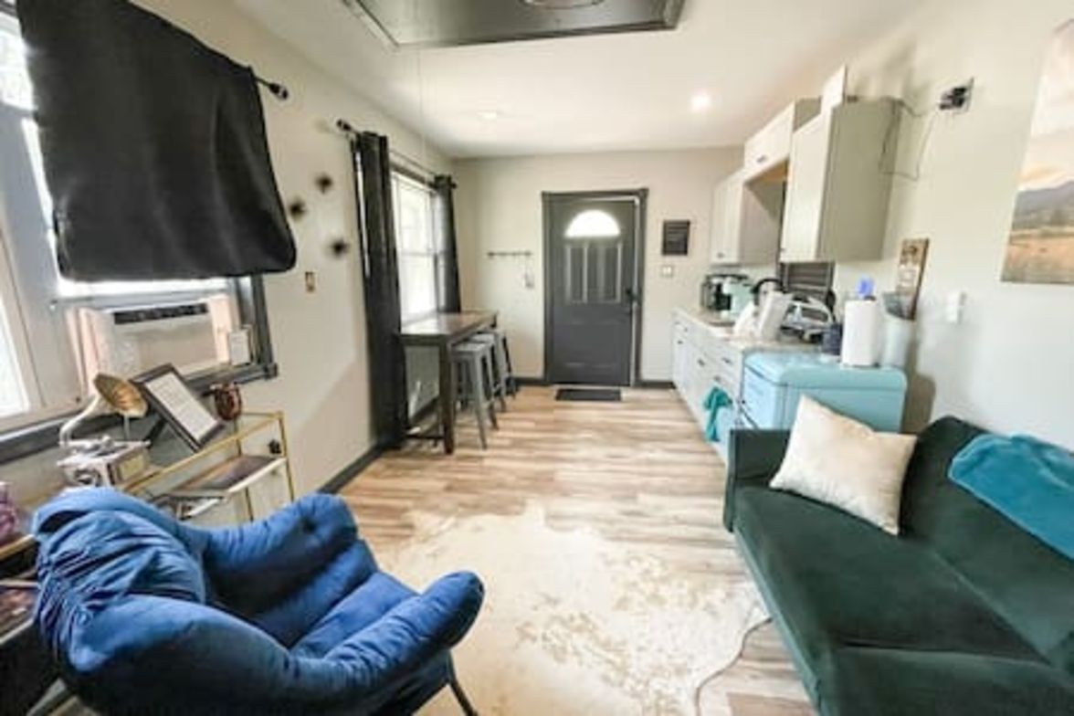 Una habitación con una silla azul, un sofá verde, una zona de cocina y una puerta que da a otra habitación.