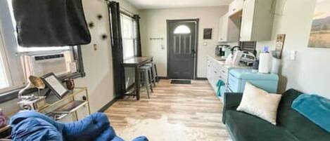 Una habitación con una silla azul, un sofá verde, una zona de cocina y una puerta que da a otra habitación.