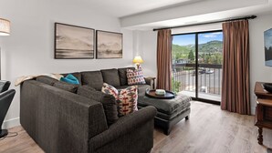 A modern living room with a sectional sofa, a small table with a lamp, and a view of a mountainous landscape through large windows. - Shadow Ridge 406 | Walk to Lifts | Mtn Views | Kitchen | Fireplace | Updated | Pool & Hot Tub (Park City)