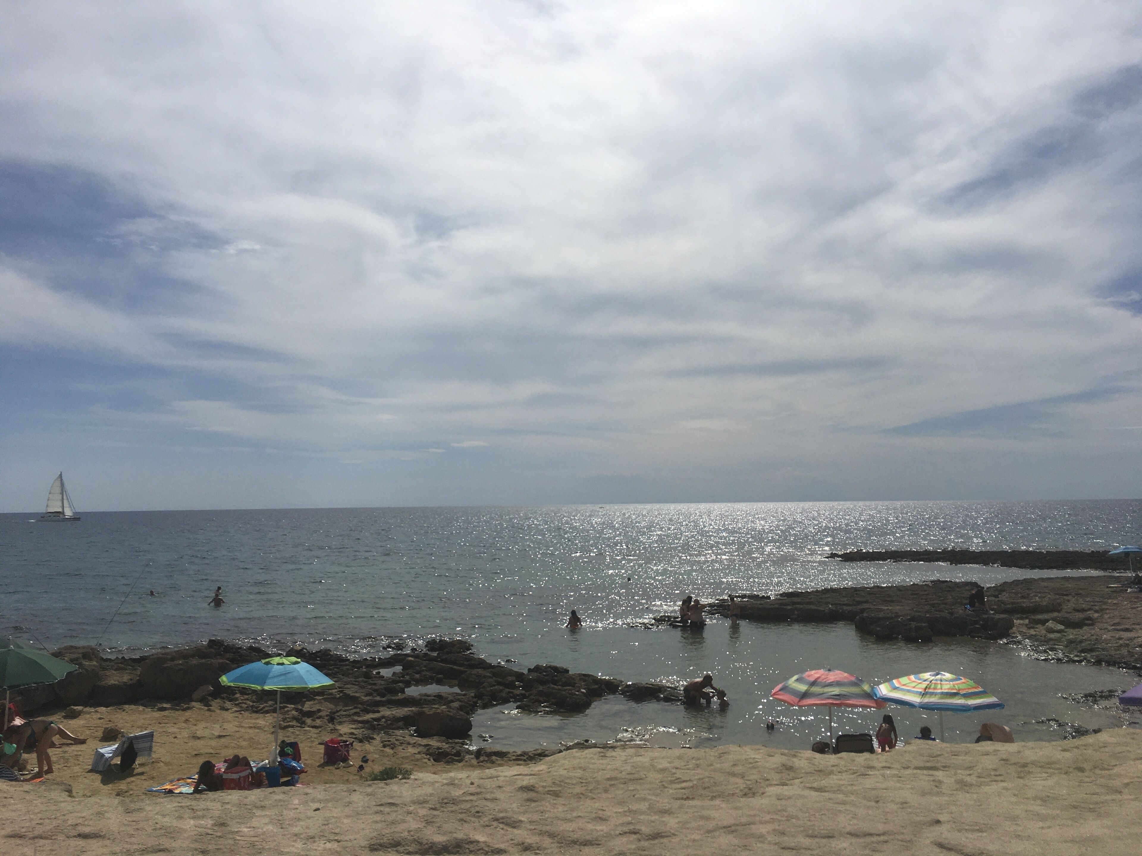 A beach with people relaxing under umbrellas, a sailboat in the distance, and rocky formations near the water.